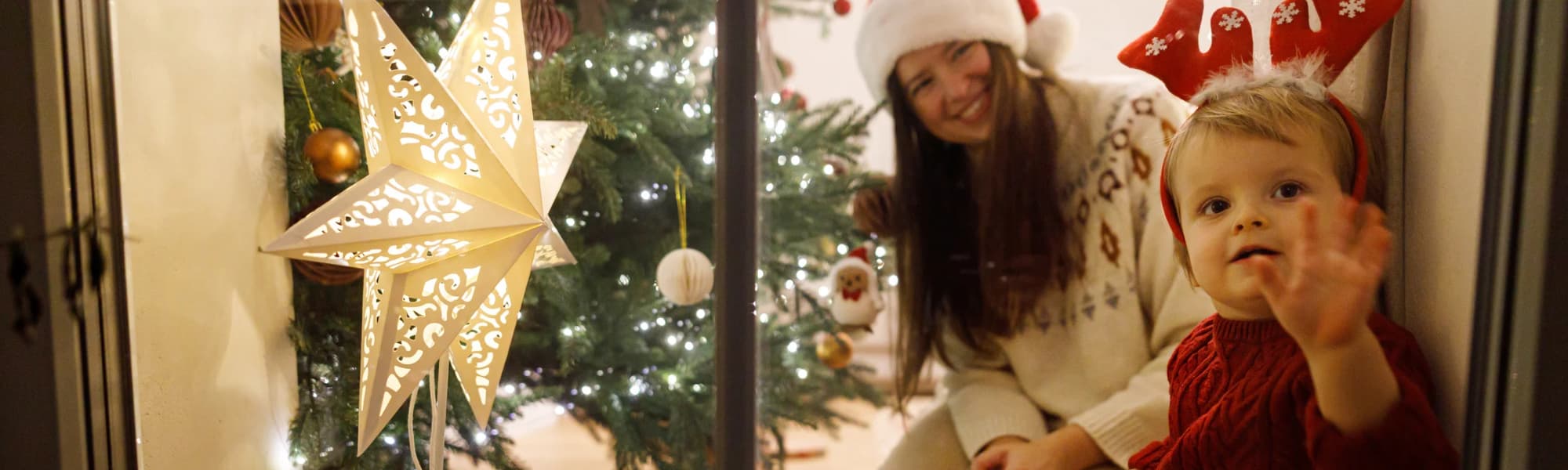 A mom and her child looking out a window with a Christmas tree behind them.